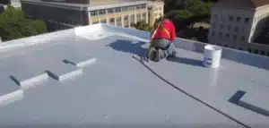 Person working on TPO roofing in San Diego with a gray coating, next to a white bucket and city buildings in the background.